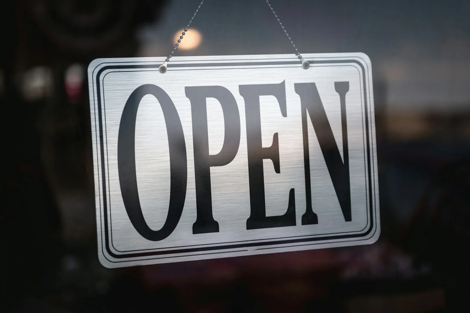 店家玻璃門上用細繩掛著一塊銀白色的「OPEN」營業告示牌，背景是模糊的室內燈光